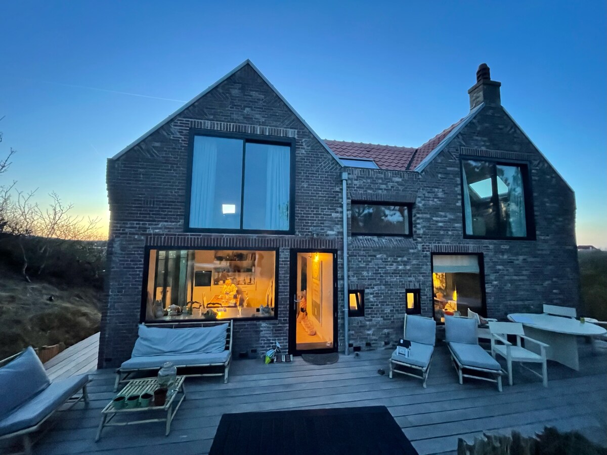 The exterior of the refurbished house is shown at dusk, with large glass windows reflecting the soft evening light. Comfortable lounge chairs are positioned on the wooden terrace, and a warm glow emanates from the interior, suggesting a cozy atmosphere inside.