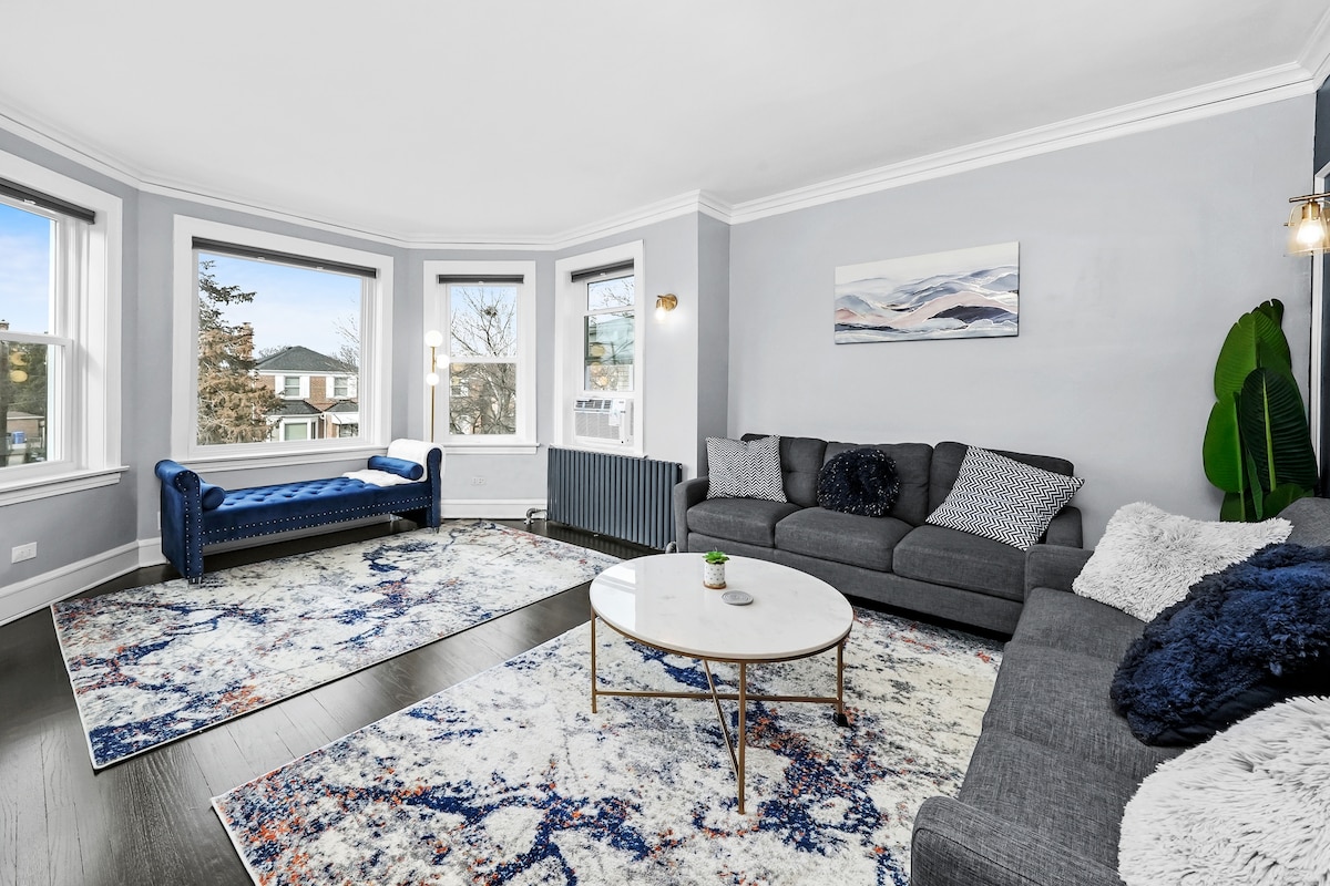 The spacious living area features large bay windows that allow ample natural light. A comfortable sectional sofa and a plush blue sofa are arranged around a coffee table. A modern rug with a colorful abstract design enhances the serene atmosphere.