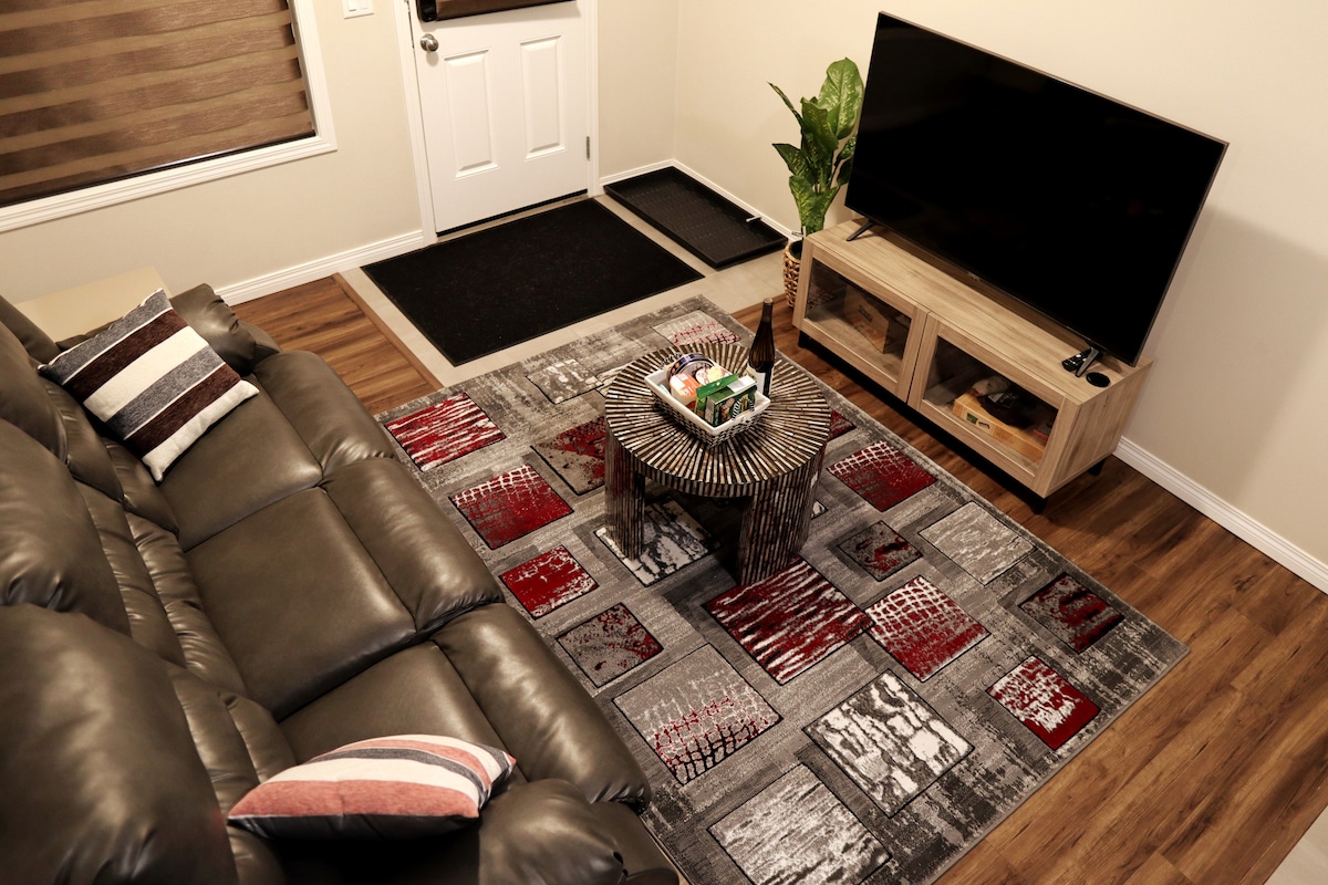 A cozy living area features a leather sofa positioned around a decorative coffee table. A modern TV stand holds a television, while a textured rug adds visual interest beneath the furniture. A green plant brings a touch of nature to the muted palette of the room.