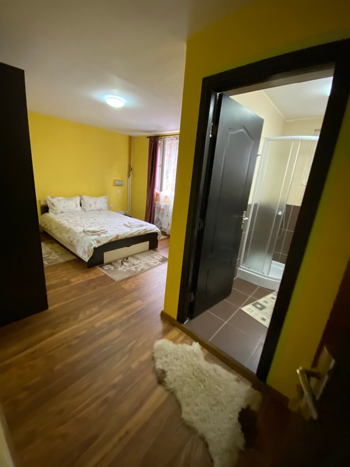 A bright bedroom features yellow walls and a spacious bed with a floral-patterned spread. Light filters through a nearby window adorned with sheer curtains. A private bathroom is visible, showcasing a modern shower stall and tiled flooring, enhancing the room's practicality.