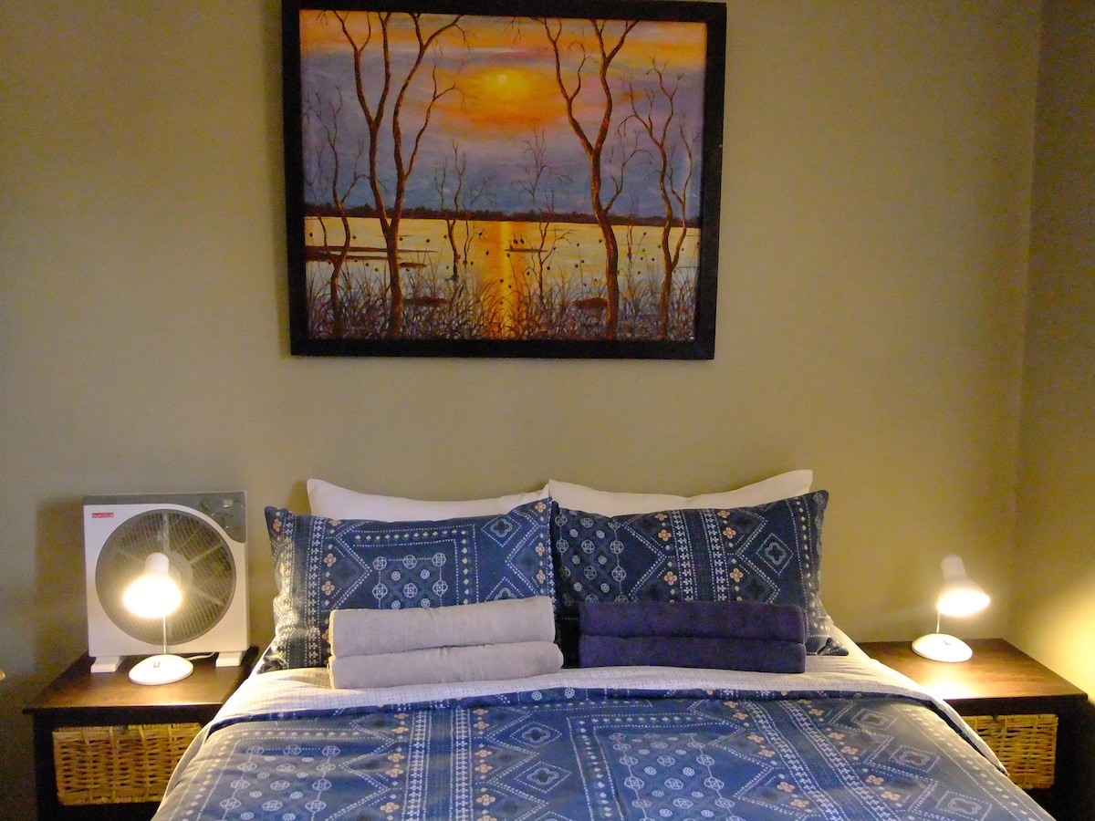 A cozy sleeping area features a spacious bed adorned with patterned blue bedding. Two bedside lamps provide soft lighting. Above the bed, a landscape painting depicts a serene sunset over water, while fresh towels are neatly arranged on the bed.