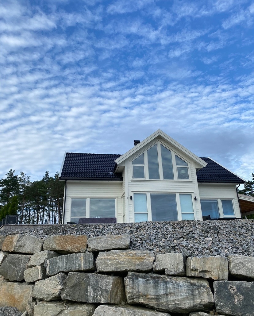 A modern house is presented with a sloped roof and large windows, allowing ample natural light. The exterior is complemented by a stone terrace, emphasizing the home's elevation. Surrounding greenery enhances the tranquil setting against a partly cloudy sky.