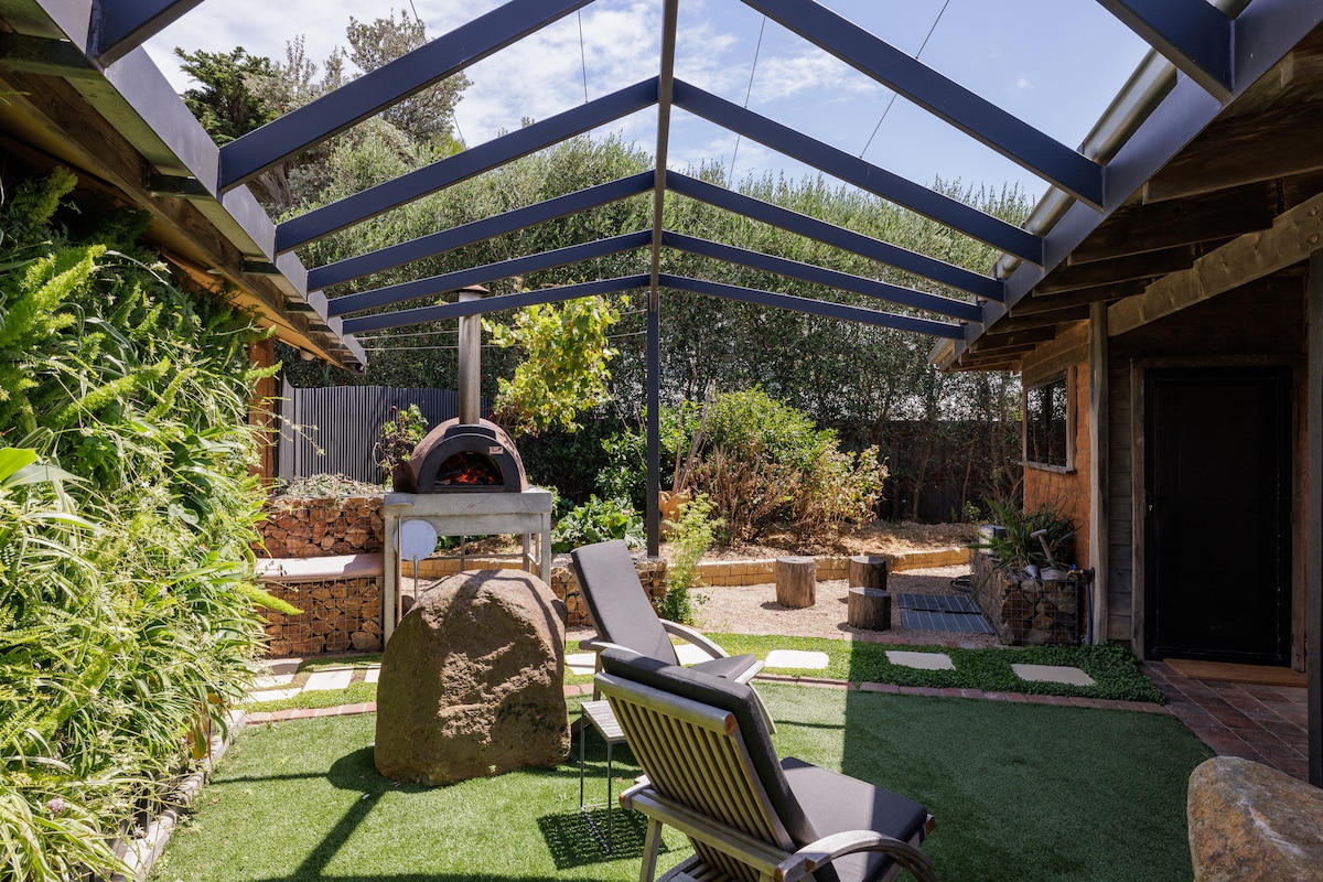 An outdoor space is showcased under a metal frame structure featuring a pizza oven and stone accents. Comfortable lounge chairs are positioned on a green surface, surrounded by lush shrubs and mature trees, creating a serene garden atmosphere.