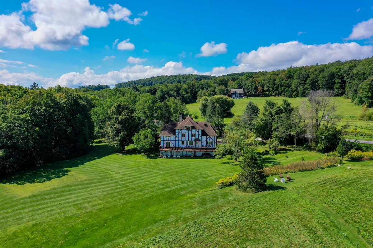Aerial view of a large estate surrounded by lush green lawns and trees, set against a backdrop of rolling hills and blue sky. The home features a mix of architectural styles with a spacious design, offering a serene and inviting outdoor space.