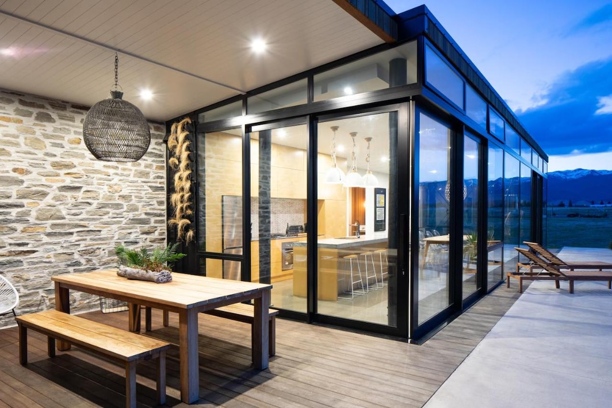 An expansive outdoor patio is featured with a large wooden dining table and chairs. Glass walls provide a view into a modern kitchen with light wood cabinetry. Soft lighting enhances the evening atmosphere, with mountains visible in the distance under an early twilight sky.
