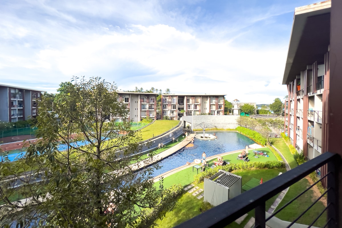 The view from the balcony offers a serene landscape featuring a landscaped pool area surrounded by green lawns. Residents are seen enjoying the sun beside the water. The buildings in the background frame the peaceful setting, enhancing the overall ambiance.