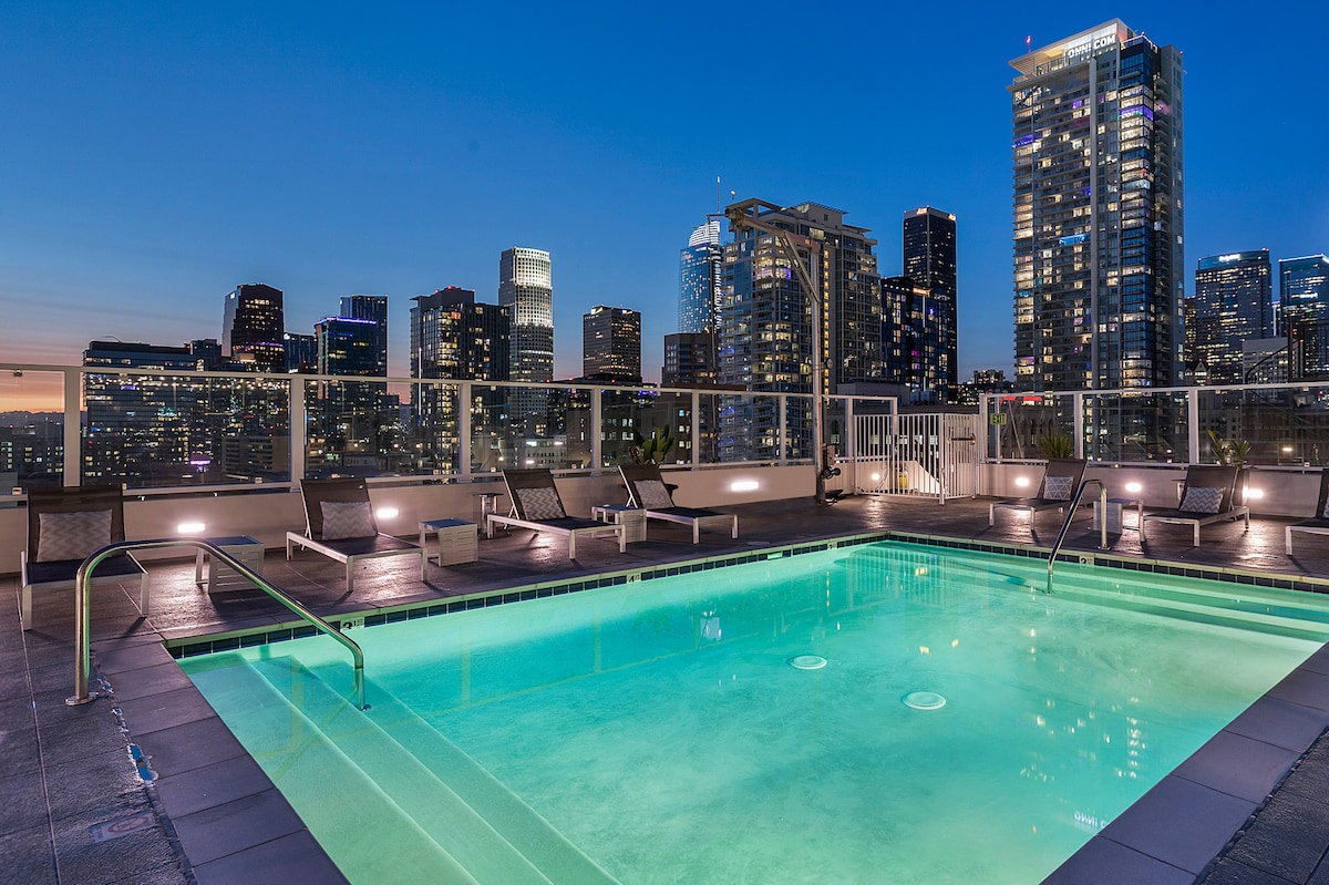 A rooftop pool area is illuminated under a twilight sky, showcasing clear water framed by modern lounge chairs. The city skyline serves as a stunning backdrop, with various architectural heights and lights reflecting the vibrant energy of downtown.