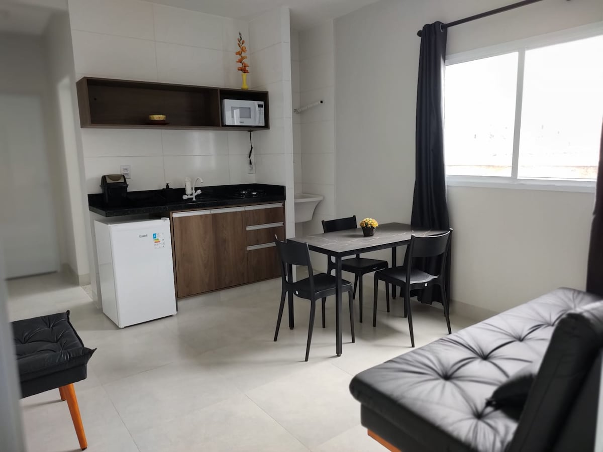 A modern kitchen area is shown, featuring a dark countertop with a microwave, sink, and refrigerator. A wooden dining table with four black chairs is positioned nearby, alongside a black sofa. Natural light flows through a large window, complemented by dark curtains.