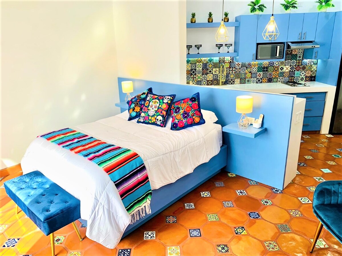 A vibrant sleeping area features a spacious bed adorned with a striped blanket and decorative cushions. Colorful wall tiles enhance the backdrop, while the kitchenette is positioned against the wall, showcasing cheerful patterned accents. Two stylish stools are placed at the foot of the bed.