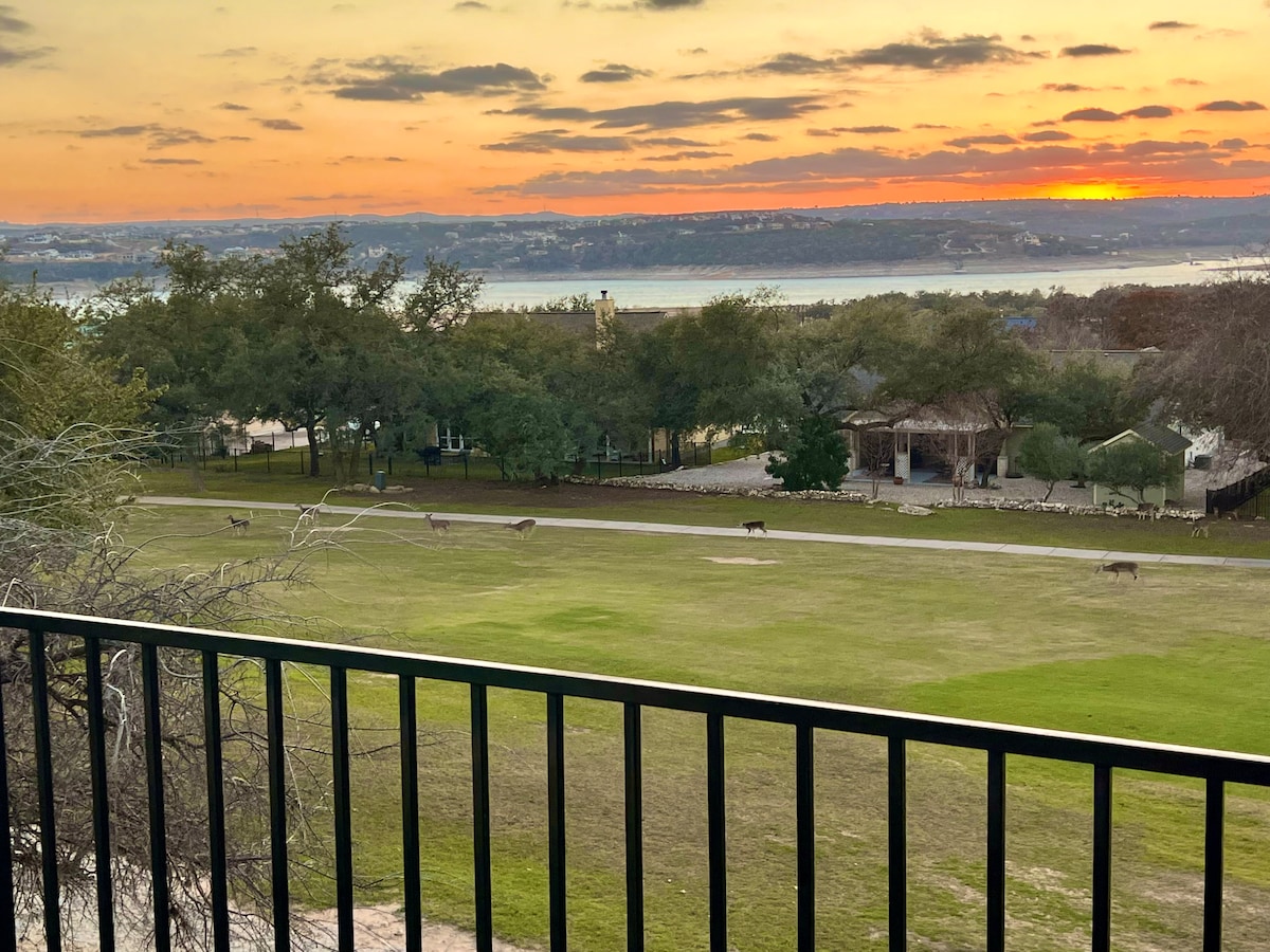 A serene sunset view showcases a grassy area framed by trees, with golfers visible in the distance. A gentle slope leads down towards Lake Travis, where the horizon is illuminated by soft hues of orange and pink. Several deer can be seen grazing peacefully.