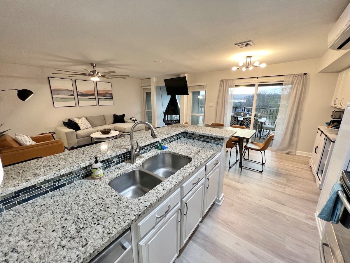 A bright, open kitchen features a granite countertop with a dual sink, positioned to provide a view of the living area. Light wood flooring extends into the dining space, where a table is set for four. Large windows allow natural light to fill the room.