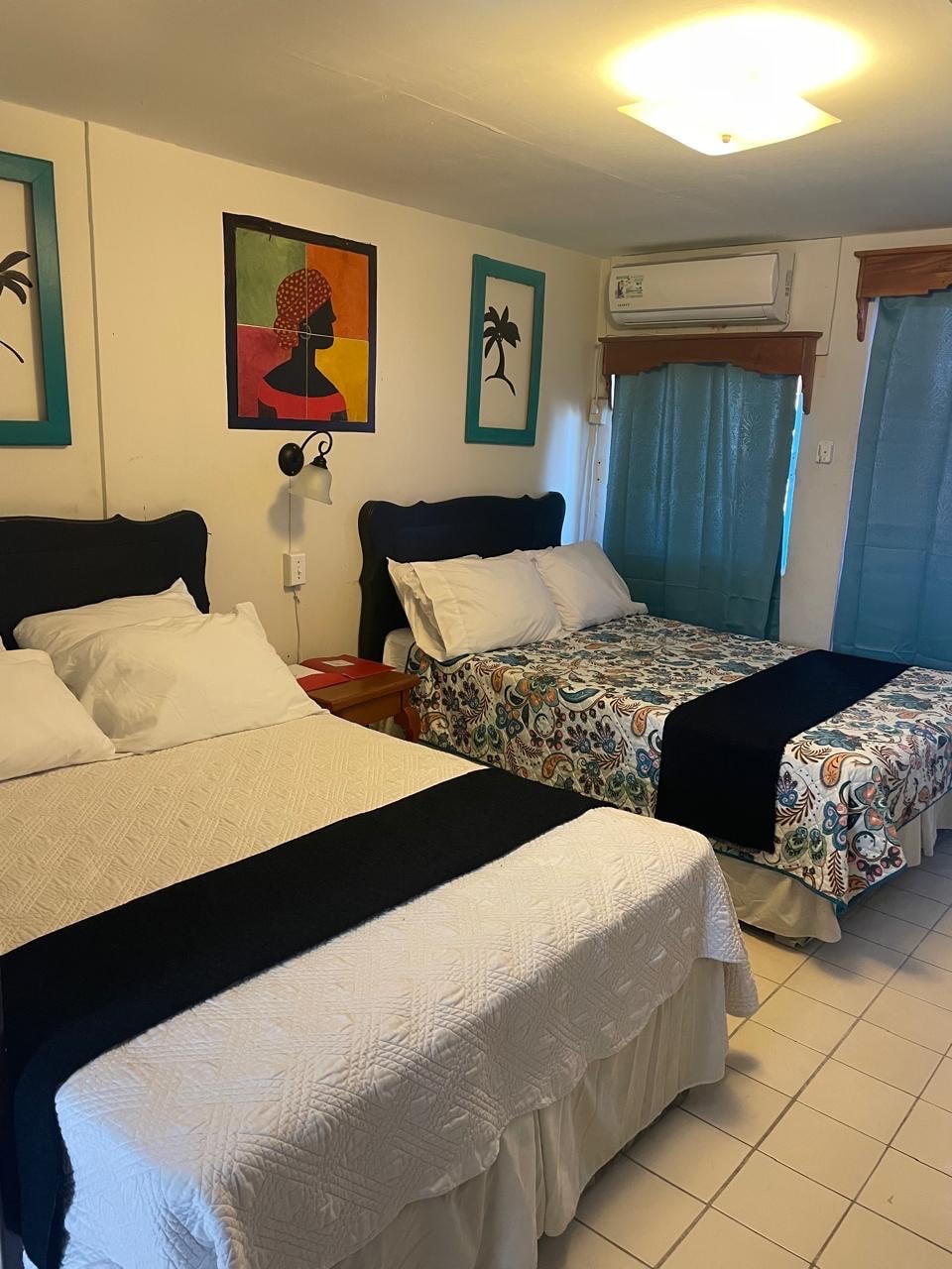 A private room features two double beds, one draped in a white quilt and the other covered with a patterned blanket. Colourful artwork adorns the wall above, complemented by teal accent frames. Window treatments allow natural light to filter in, enhancing the room's airy feel.