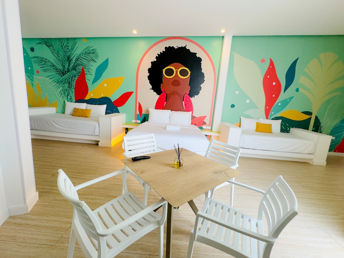 An inviting lounge area features two spacious white sofas positioned against a colorful mural with tropical themes. A light wooden table is surrounded by four white chairs, creating a perfect space for relaxation and socializing.