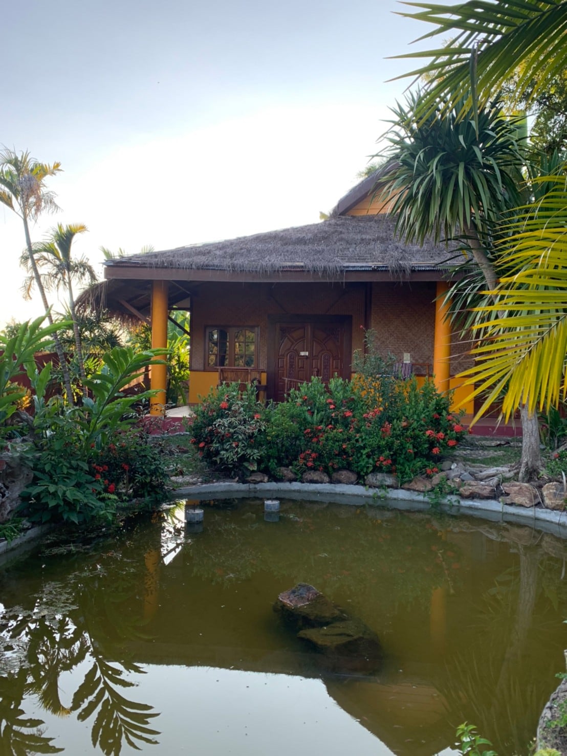 A charming building with a thatched roof is surrounded by lush greenery and colorful flowers. A tranquil pond reflects the structure, while tropical plants frame the entrance, enhancing the natural setting. The peaceful atmosphere invites relaxation and connection with nature.