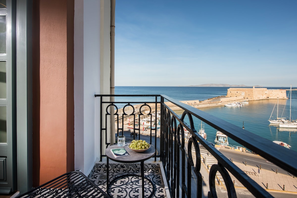 A private balcony is shown, featuring a small round table with a bowl of grapes on it. The view reveals the Venetian Harbour and Koules fortress, with boats visible in the water and a clear blue sky overhead.