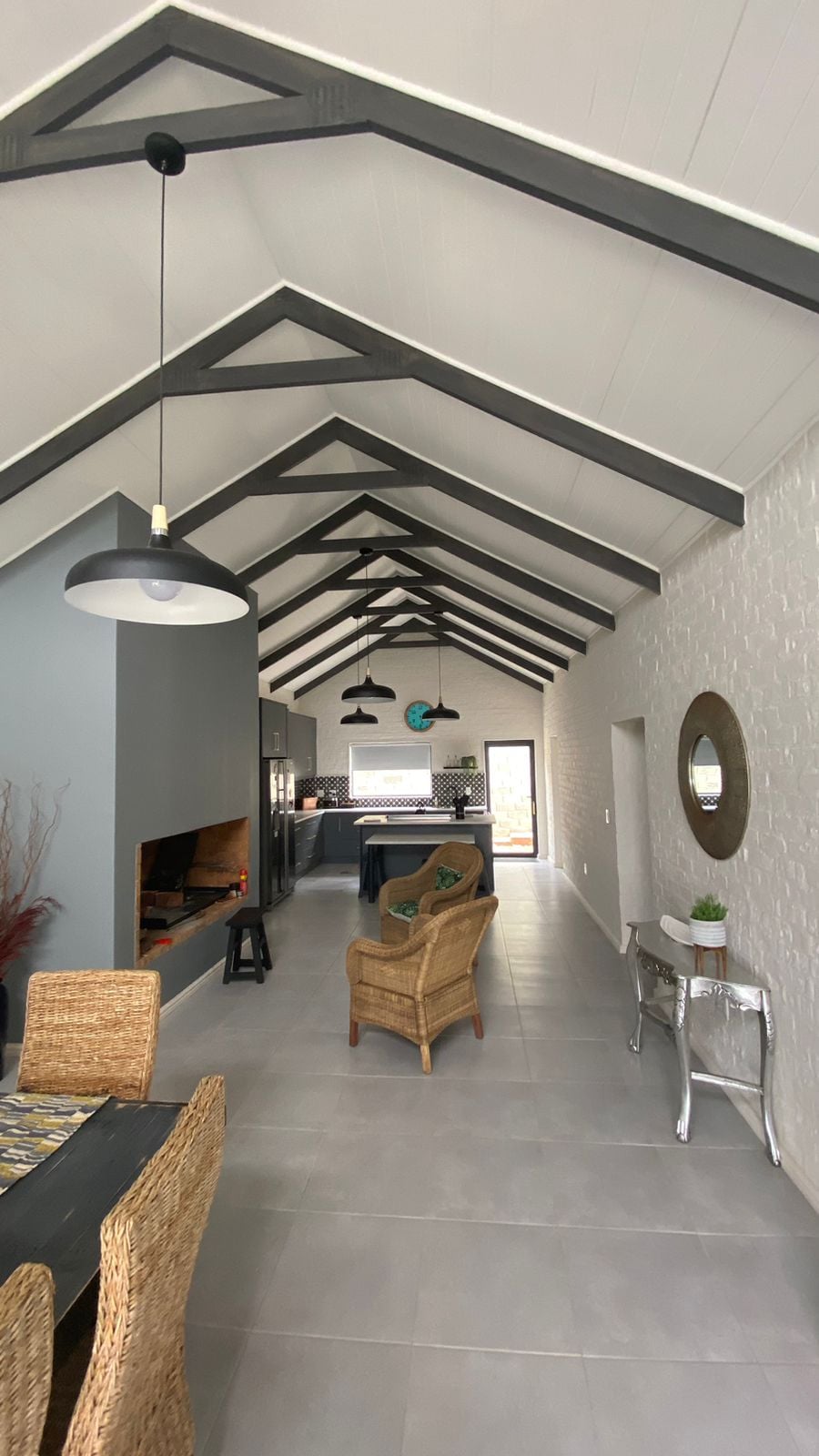 The open-plan living area features high, vaulted ceilings adorned with wooden beams. Natural light floods the space, illuminating a cozy lounge area and a dining space. A modern kitchen is visible, and a decorative mirror is placed on the wall, enhancing the ambiance.