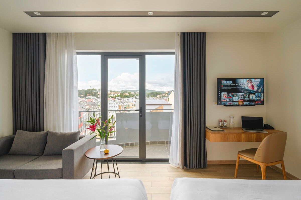 A bright living space features large glass doors opening to a private balcony, offering views of the cityscape. A gray sofa is positioned beside a small table, while a wooden desk displays a Smart TV and a few amenities. Light curtains frame the windows, enhancing the airy ambiance.