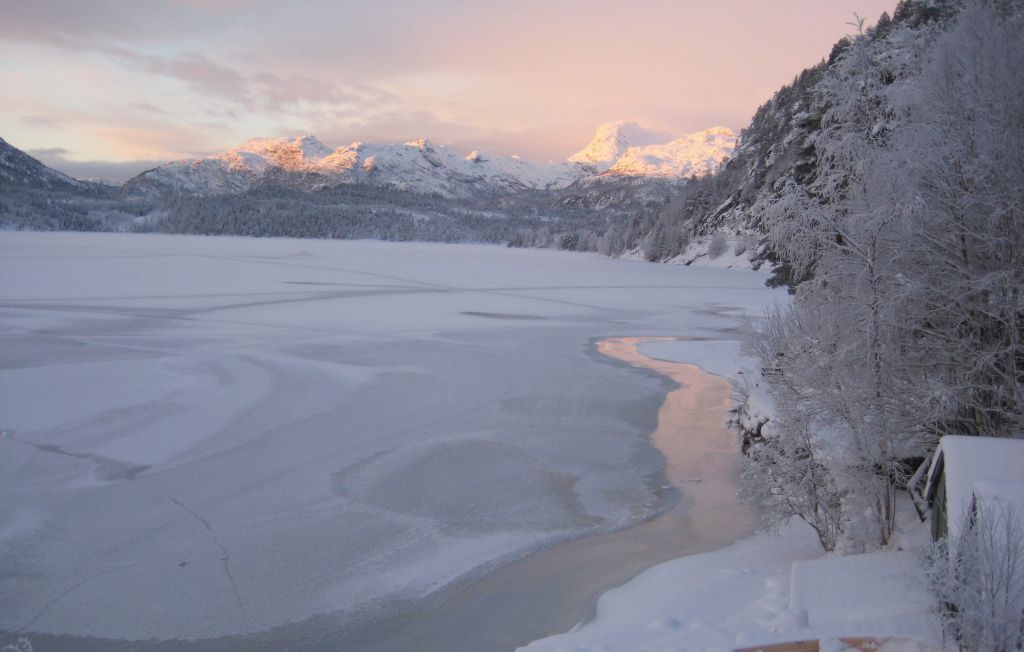 A serene winter landscape is depicted, featuring a frozen lake surrounded by snow-covered mountains. Soft pastel colors from the sunset illuminate the scene. Frosted trees line the shore, and the stillness of the ice creates a tranquil atmosphere.