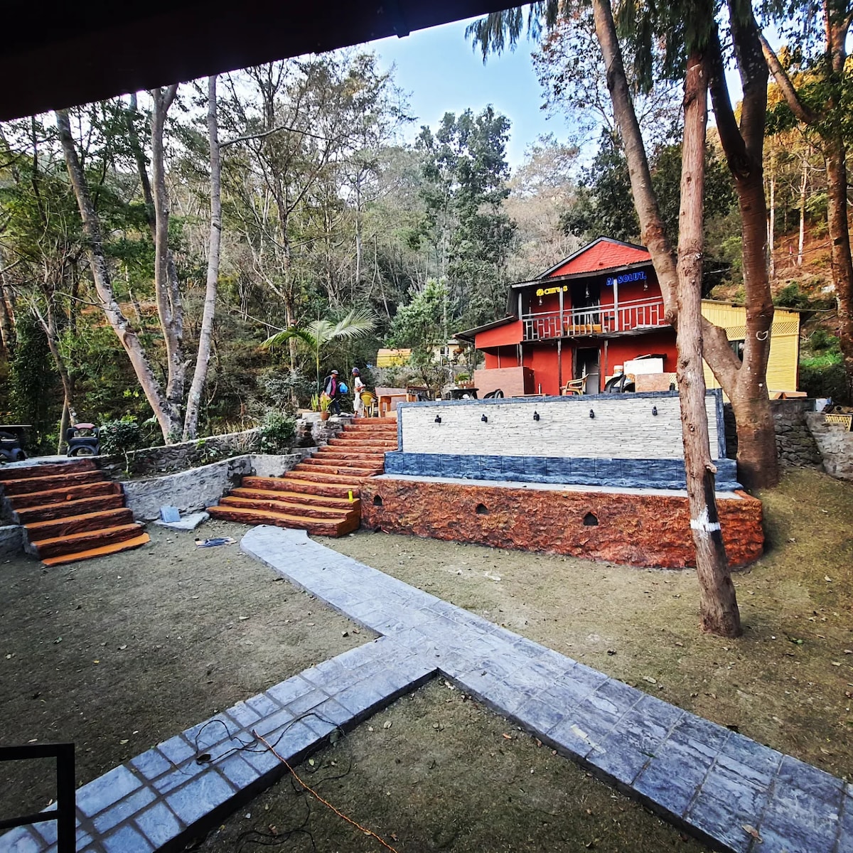 The outdoor area features landscaped pathways and multiple levels of terraced gardens leading to a two-story building. Steps ascend to a patio area with stone walls and water features, surrounded by trees and greenery, offering a natural and serene environment.