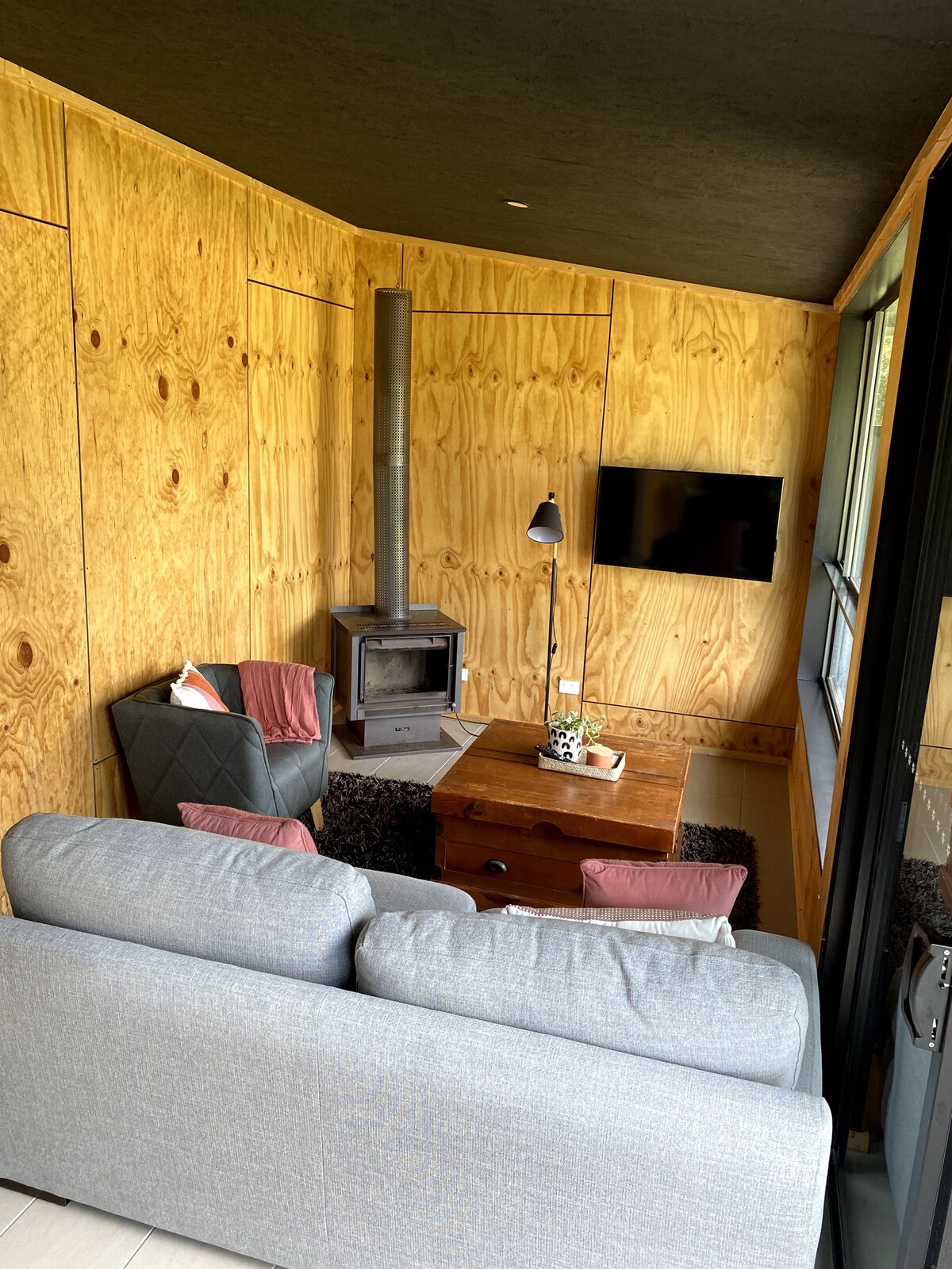 A cozy sitting area features a comfortable gray sofa facing a wooden coffee table. Two armchairs add to the seating options. A wood-burning stove is positioned beside a wall, and a flat-screen TV is mounted nearby. Natural light filters through large windows.