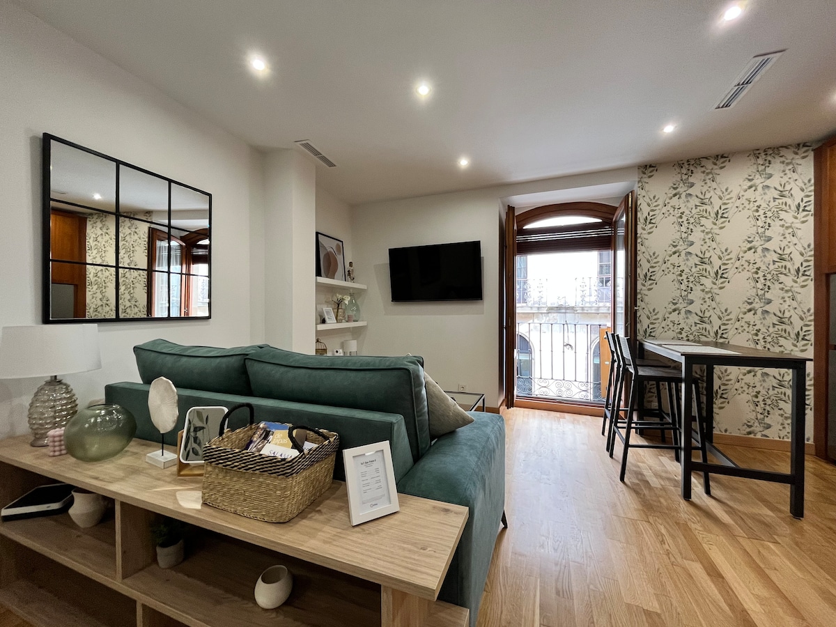 A cozy living area features a light green sofa facing a wall-mounted television. Natural light floods in through large windows with wooden frames. A wooden shelf displays decorative items, and a dining table set for two is present in the corner, complemented by modern black chairs.