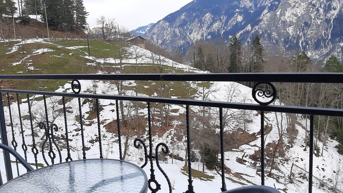 A balcony view showcases a landscape of snow-covered ground and distant mountains. A round glass-top table is seen against an intricate metal railing, with greenery and trees partially visible in the background.