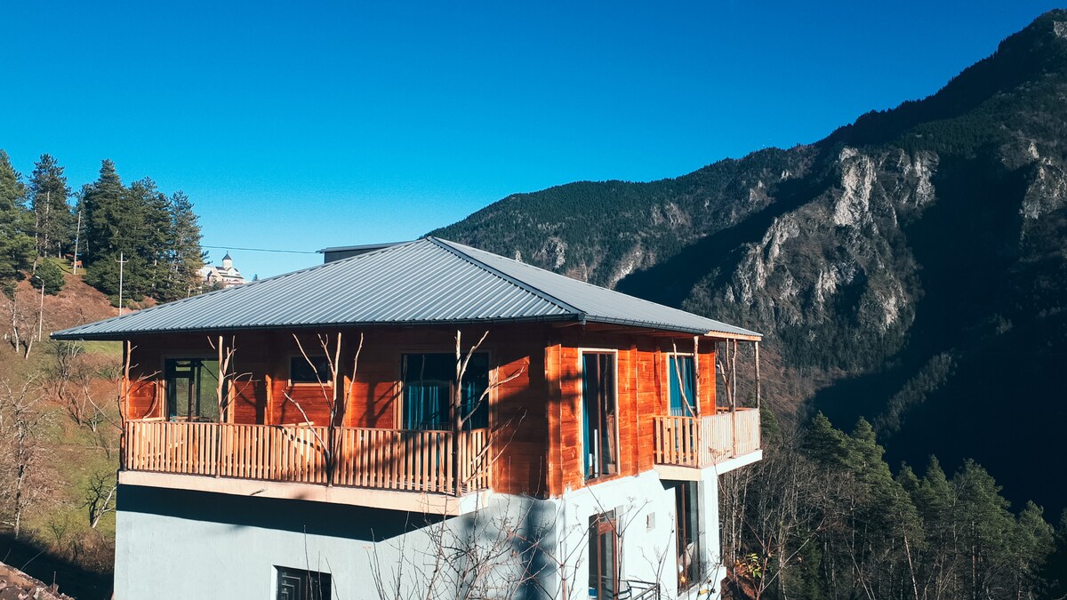 The exterior of a two-story wooden building is shown, featuring large windows and a wrap-around balcony. Surrounded by mountainous scenery, the structure's sloped roof complements the landscape, with clear blue skies in the background enhancing the natural setting.