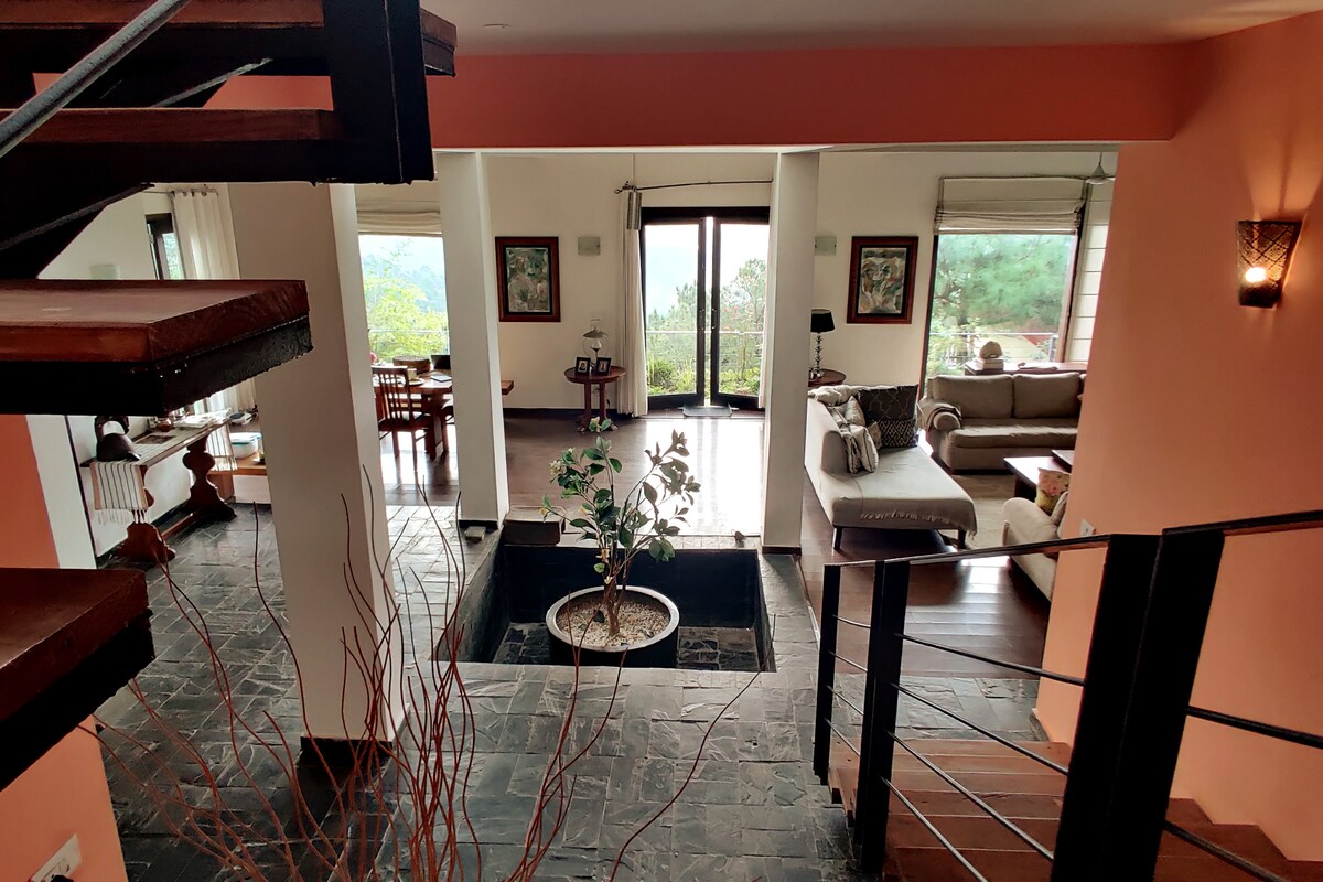 The interior showcases an open living space with large windows providing hillside views. A central indoor garden element is framed by a spiral staircase. The carefully arranged seating areas invite relaxation, while dining furniture is visible in the background, creating a cohesive and spacious layout.