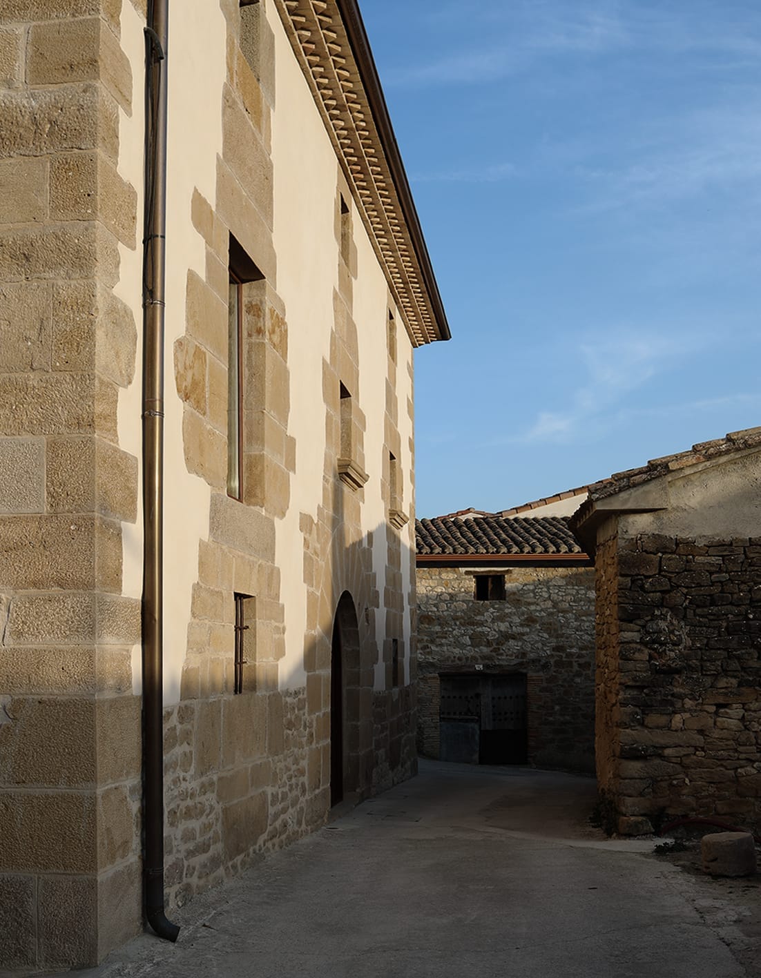 The image captures the exterior of a historic building, showcasing its sturdy stone walls and arched doorway. Soft shadows are cast along the façade, and a clear blue sky is visible above, highlighting the architectural details and the structure's integration within its surroundings.