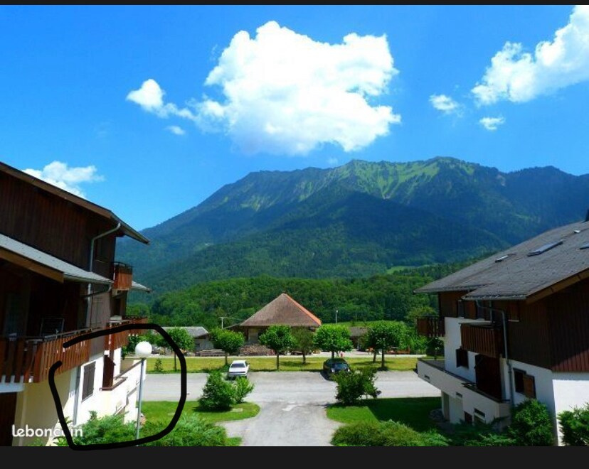 The image captures a scenic view featuring multiple residential buildings nestled among lush greenery, with a prominent mountain in the background. Bright blue skies and fluffy clouds create a vibrant atmosphere, while trees and well-maintained pathways enhance the overall setting.