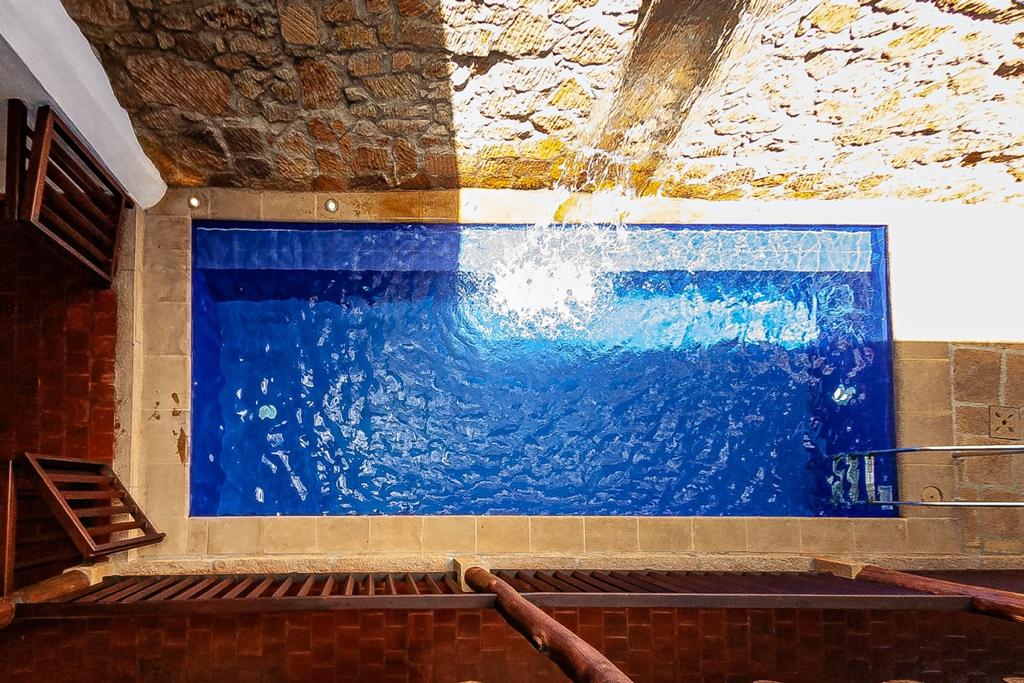 A modern swimming pool features clear blue water, reflecting sunlight. The surrounding stone walls add texture, while the pool's edge is lined with tiles. A gentle waterfall can be seen, enhancing the serene environment.