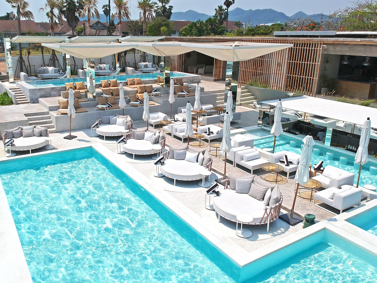 An expansive pool area features multiple elegant lounge chairs and daybeds, all arranged around a bright, turquoise infinity pool. Sheltered seating areas with umbrellas provide comfort, while modern furnishings enhance the overall aesthetic. Lush greenery and palm trees are visible in the background.