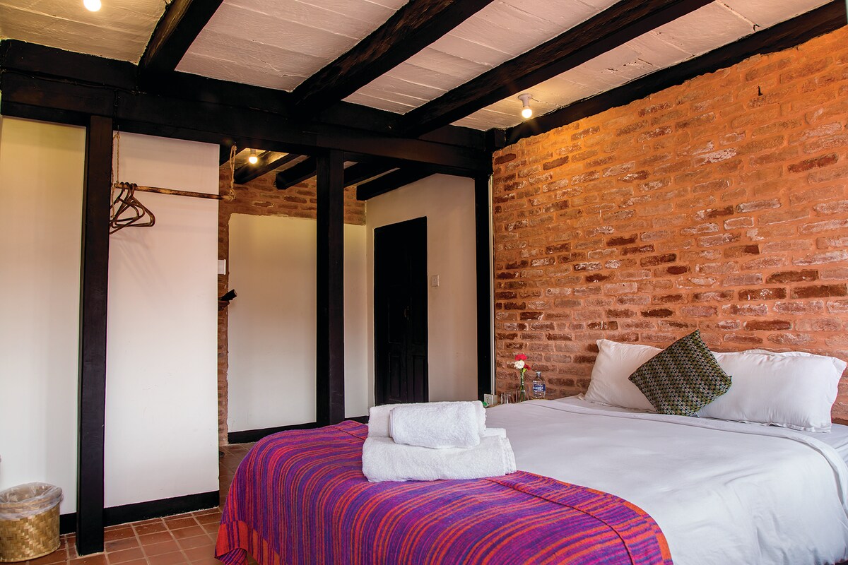 A cozy sleeping area features exposed brick walls and wooden beams. A spacious bed is adorned with a colorful blanket, accompanied by neatly arranged towels. A small flower adds a touch of warmth to the space, which is illuminated by soft overhead lighting.