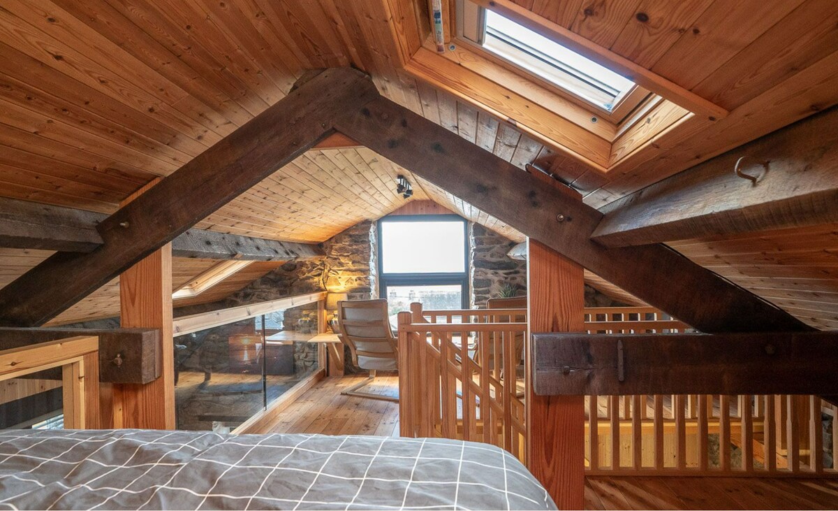 An attic space features wooden beams and a sloped ceiling, lending a rustic feel. A skylight allows natural light to flood the area, illuminating the wooden floors and walls. A cozy corner chair is positioned near the railing, offering views of the surroundings.
