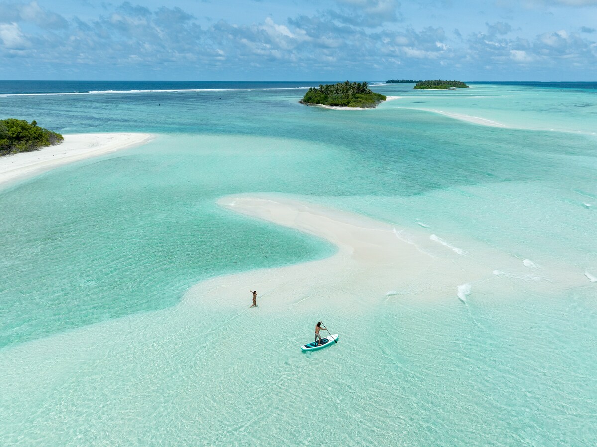 A serene landscape is captured, featuring shallow turquoise waters and white sandy shores. A lone paddleboarder glides through the gentle waves, with lush green islands visible in the distance under a partly cloudy sky.