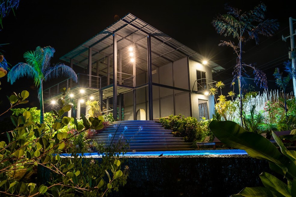 The house is illuminated at night, showcasing a modern structure with large windows and an open design. Surrounding foliage adds to the ambiance, while a swimming pool reflects shimmering lights in the foreground. The setting highlights the natural beauty of the environment.
