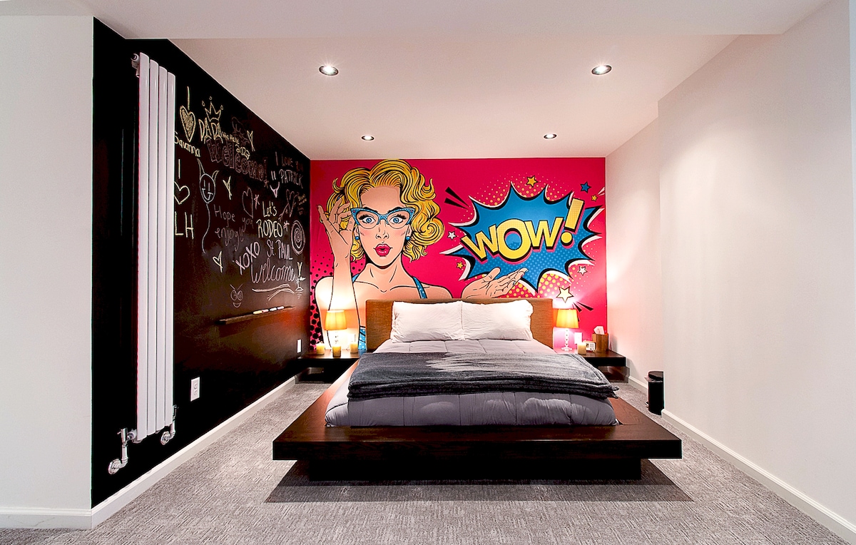 A modern bedroom features a spacious bed with a dark wood frame and luxury linens. A vibrant pop art mural with a 'WOW!' graphic serves as a focal point. Soft lighting fixtures complement the minimalist design, while a black chalkboard wall invites creativity.