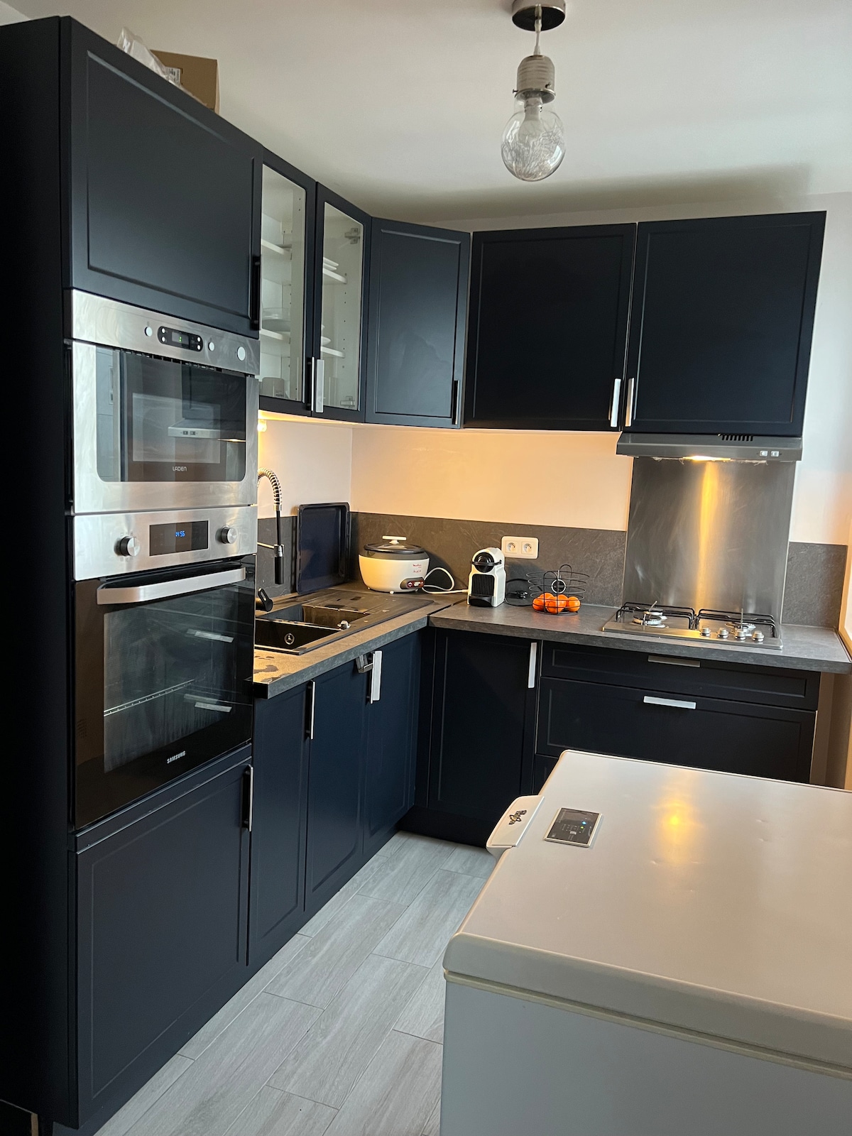 A modern kitchen is displayed with dark cabinetry and stainless steel appliances. An oven and microwave are built into the cabinetry, while a stove is positioned beneath a sleek extractor hood. Ample counter space is available, and the area is well-lit, enhancing functionality.