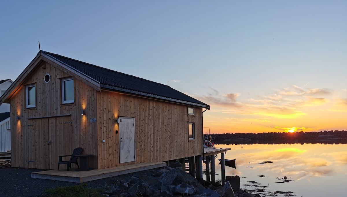 A newly constructed wooden cabin is situated by the water's edge, with large windows reflecting the colorful sunset. The exterior features warm wooden tones and subtle lighting. A seating area is visible on the porch, overlooking the serene water and distant shoreline.