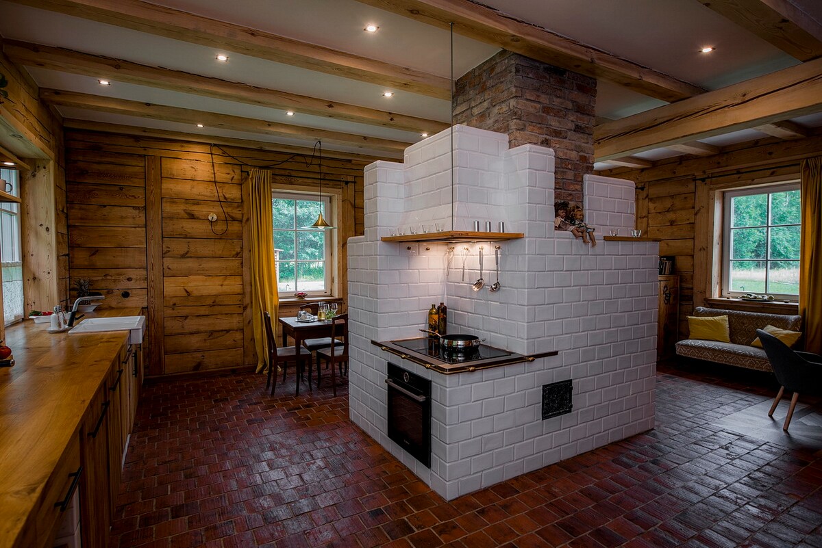The open kitchen features a central cooking area with a tiled stove, surrounded by wooden cabinetry and rustic brick flooring. Large windows provide natural light, illuminating a dining area with a table and chairs in the background, alongside a cozy seating arrangement.