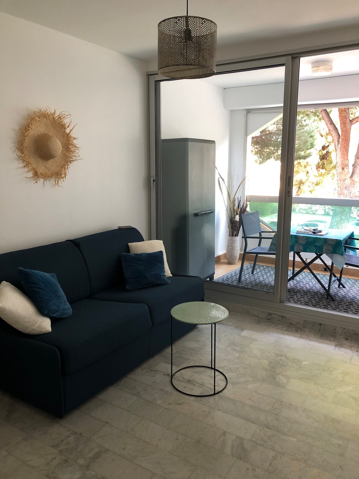 A cozy seating area features a dark blue convertible sofa adorned with light-colored cushions. A small round table is placed beside the sofa. Floor-to-ceiling glass doors lead to an outdoor loggia, visible beyond a patterned rug and a set of chairs and table.
