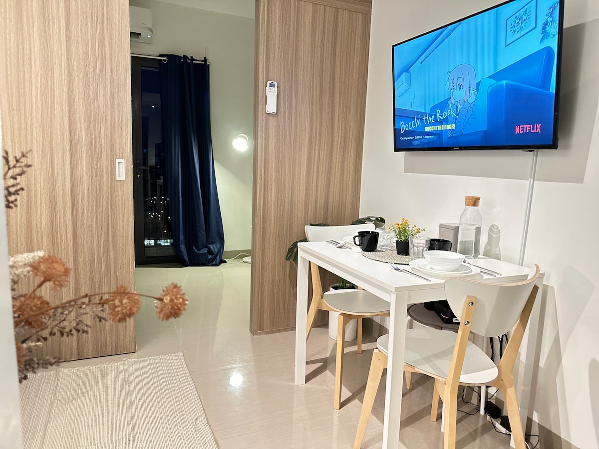 A dining area is seen with a modern white table and two chairs, set with dishes and cups. A sleek television mounted on the wall displays Netflix. A cozy corner lamp provides soft lighting, while blue curtains are drawn to reveal a view outside.