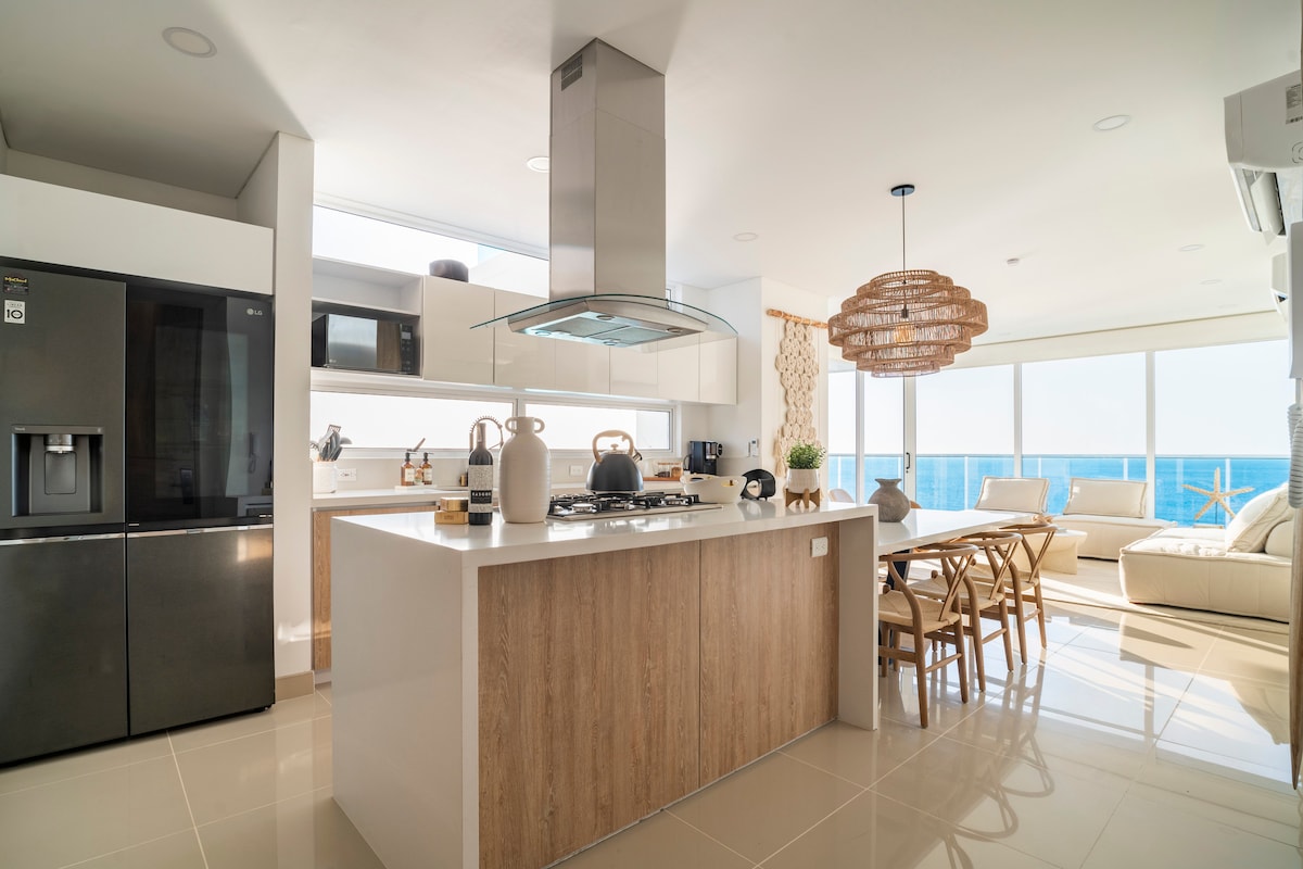 The modern kitchen features stainless steel appliances and a large island with a countertop. The dining area includes a wooden table surrounded by chairs, while floor-to-ceiling windows highlight views of the Caribbean Sea, allowing natural light to brighten the space.