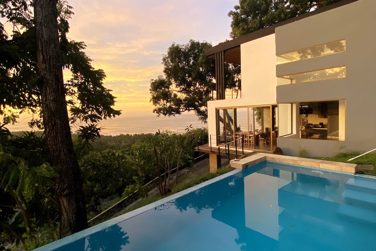 A modern villa is depicted beside a private infinity pool, surrounded by lush greenery. The large glass windows reveal a spacious interior while sunset colors reflect on the water's surface, creating a serene atmosphere. Views of the Pacific Ocean are captured in the background.