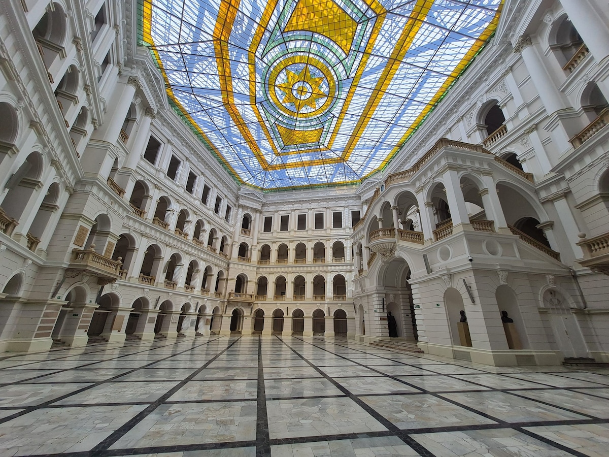 An elegant indoor space features a high glass ceiling with a vibrant stained-glass centerpiece, allowing natural light to fill the area. The marble floor displays a checkerboard pattern, while ornate archways and columns frame the view, creating a harmonious architectural environment.