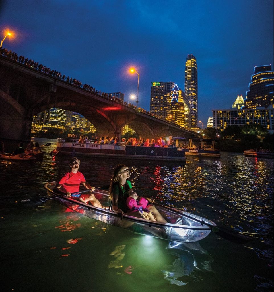A kayak glides gently through the water at night, illuminated by soft light. The skyline of Austin is visible in the background, with skyscrapers and lights reflecting on the water's surface. A large crowd can be seen enjoying the evening on the nearby bridge.