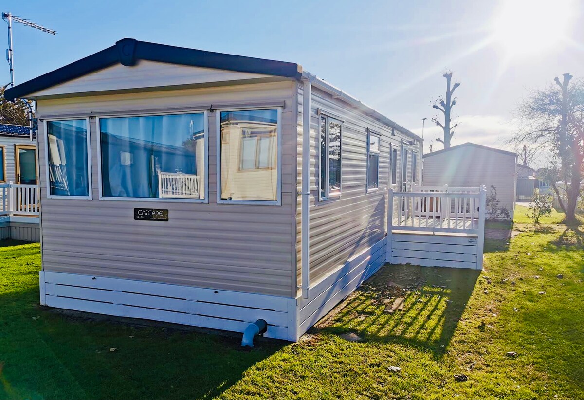 The exterior of a mobile home is depicted, featuring a light-colored façade with large windows allowing natural light in. A small deck is accessible from the side, with a grassy area surrounding the home. The setting is illuminated by bright sunlight.