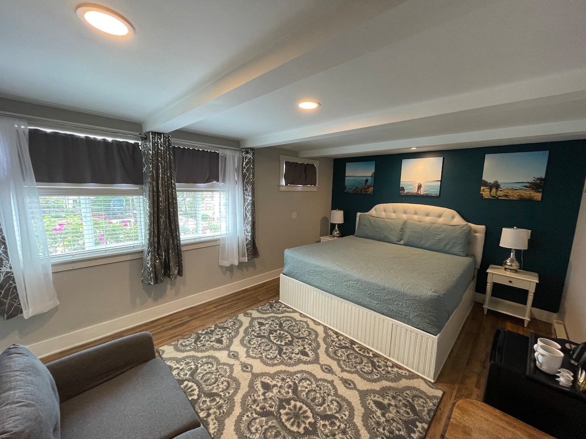 A spacious room features a king-size bed dressed in soft blue linens. Light filters through grey window treatments, highlighting a cozy seating area with a grey chair. Decorative artwork adorns the accent wall, while bedside tables with lamps add functionality.
