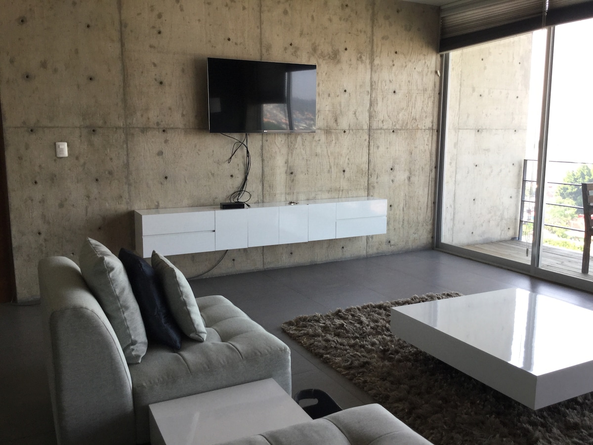 A spacious living area features a light-colored sectional sofa paired with decorative cushions. A modern coffee table sits on a textured rug. A wall-mounted flat-screen television is positioned above a minimalist white console. Large glass doors lead to a balcony, providing natural light and outdoor views.