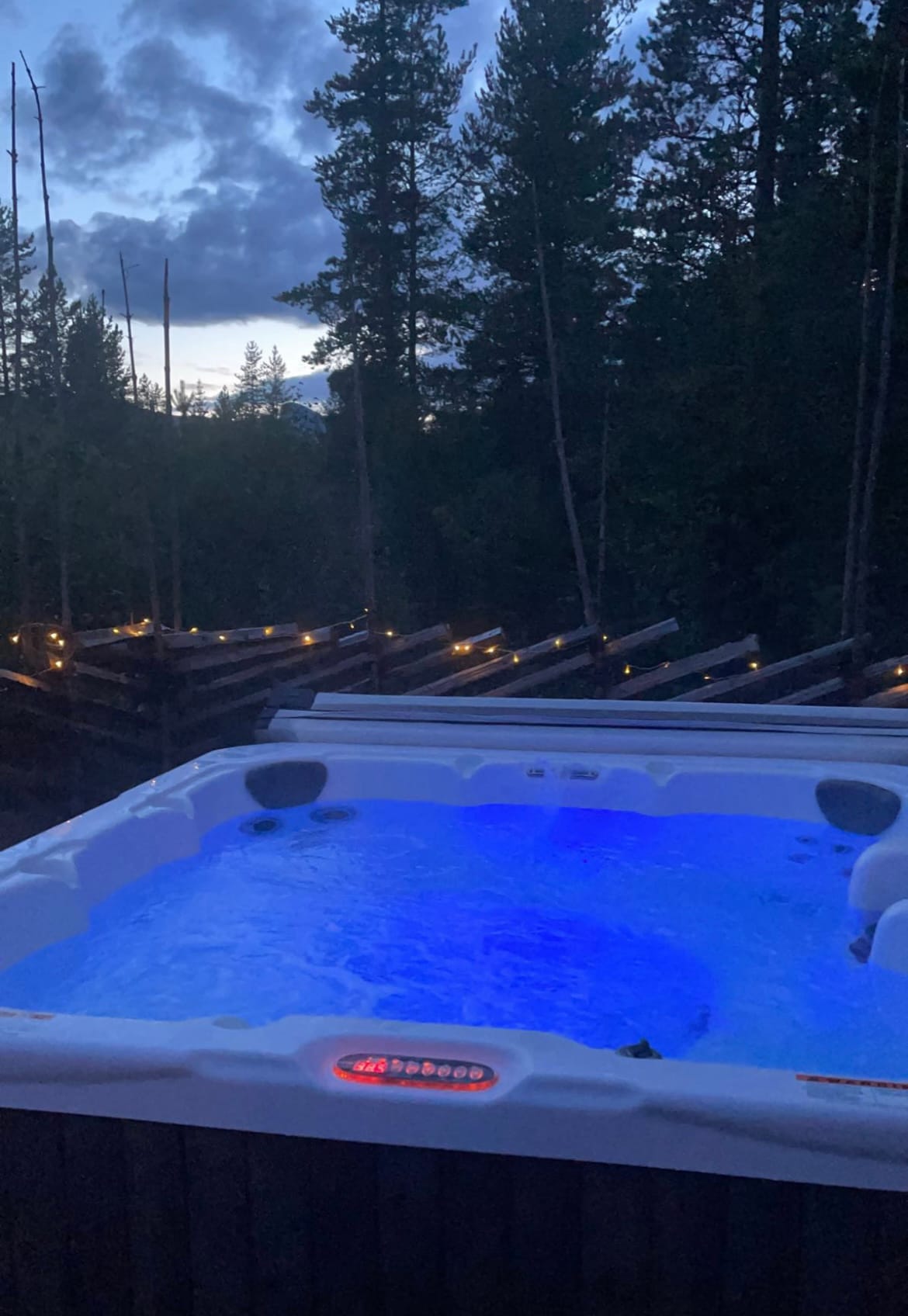 The outdoor hot tub is illuminated with soft blue lighting, surrounded by a serene forest setting. Wooden decking is visible in the background, with warm fairy lights adding a gentle glow as dusk approaches.
